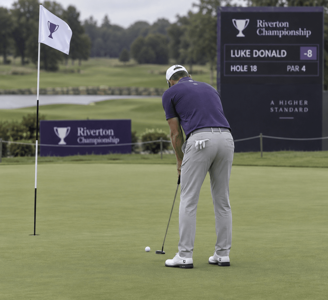Golfer Luke Donald lines up a final putt beside the flagstick on the 18th green, with a tournament scoreboard behind him.