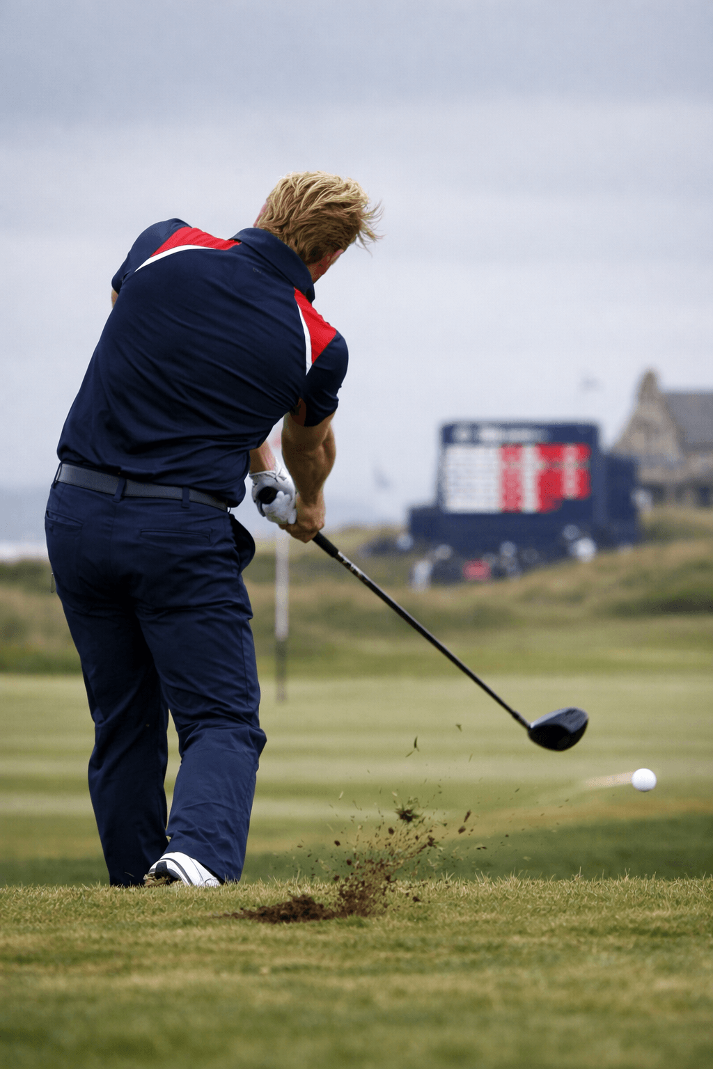 Tommy Fleetwood golfer mid-swing on a windy links, driving a ball over striped fairway turf.